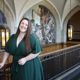 Kasee Arnett  - Lady standing with long brown hair in green dress in historic building next to railing with lights behind her